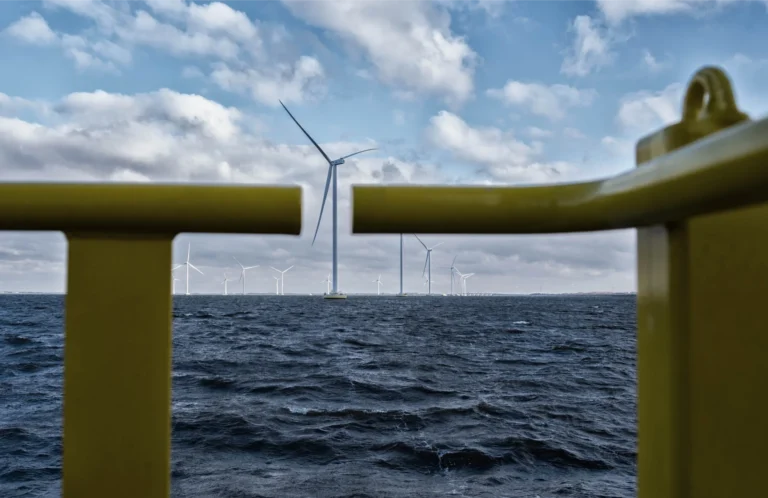 A view of offshore wind turbines generating renewable energy in the ocean under a cloudy sky, seen through the yellow railing of a nearby structure. The turbines are arranged in the distance, illustrating the scale of offshore wind energy production.