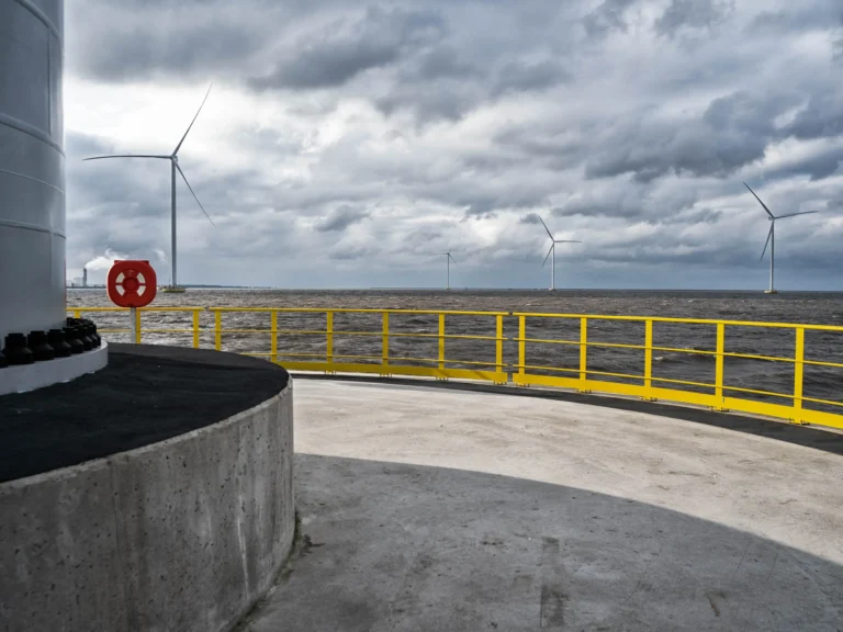 Offshore wind turbines generating renewable energy in the ocean on a cloudy day, viewed from a wind farm platform with yellow safety railings. The image highlights clean energy technology and the growing trend of offshore wind power.