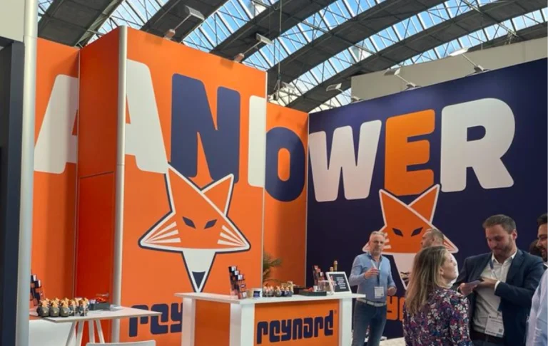 Reynard's exhibition booth at the Offshore Energy Exhibition & Conference 2024, featuring bold orange and navy blue walls with large 'POWER' lettering and Reynard's fox logo. Visitors are seen interacting around the booth, with branded displays and product samples visible on the counters.