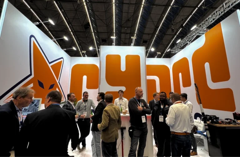 Busy trade show booth with a prominent orange logo in the background, where people are networking and having conversations under professional lighting