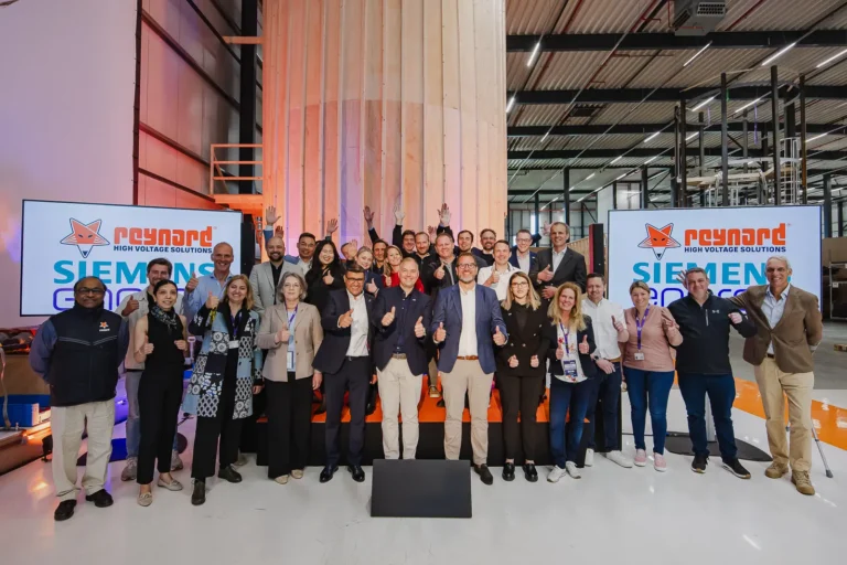 Large group photo of Siemens Energy and Reynard High Voltage Solutions teams giving thumbs up in industrial facility with company logos displayed on screens behind them