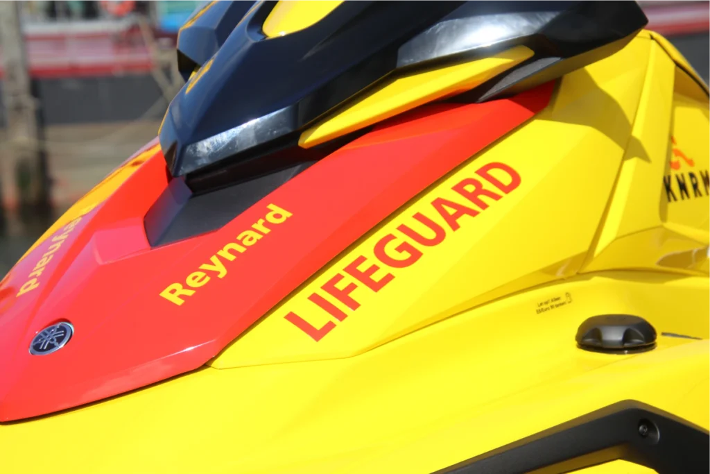 Close-up detail of yellow and red KNRM lifeguard jet ski showing Reynard company branding and LIFEGUARD text on the hull of the donated rescue watercraft